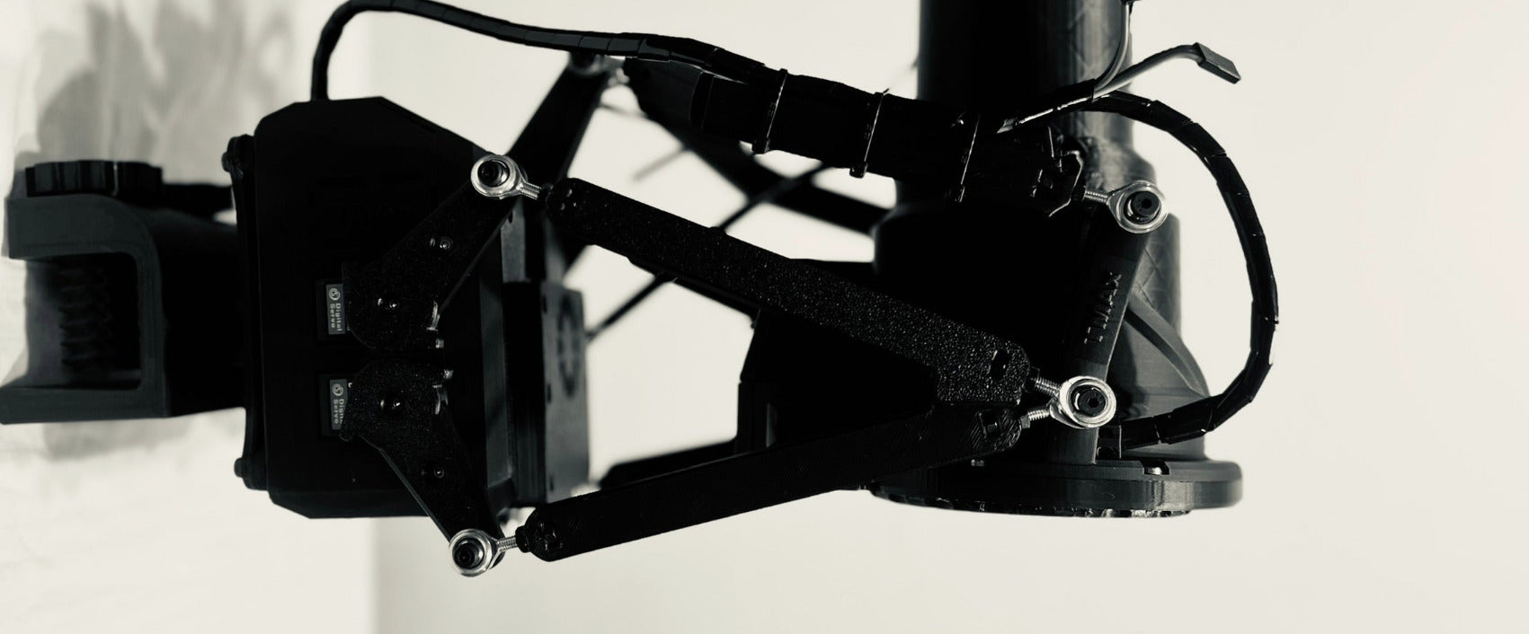 A close-up of a black camera mounting bracket or rig component with visible screws, straps, and a cylindrical attachment, photographed against a light neutral background.